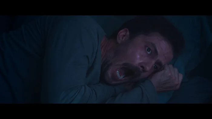 A terrified man clutching his sheets in a red-lit bedroom