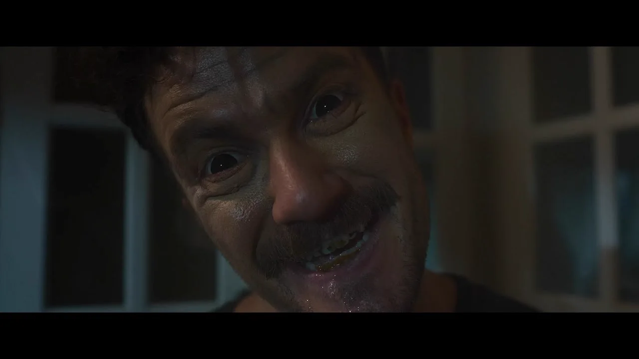 Trailer still — a crazed grinning man stares into the camera in a dark room